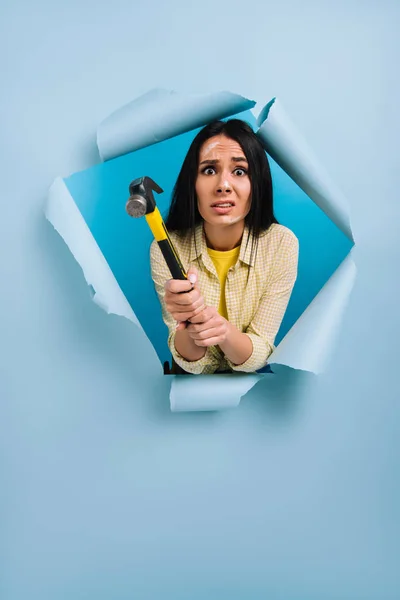 Mulher assustada trabalhador manual com rosto sujo na pintura segurando martelo em papel rasgado, isolado em azul — Fotografia de Stock