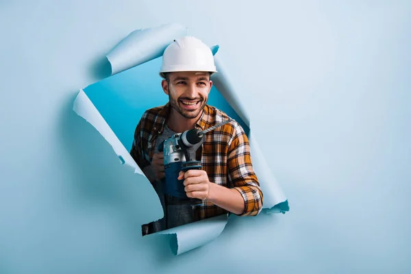 Manitas sonrientes en casco sosteniendo taladro eléctrico en papel roto, aislado en azul - foto de stock