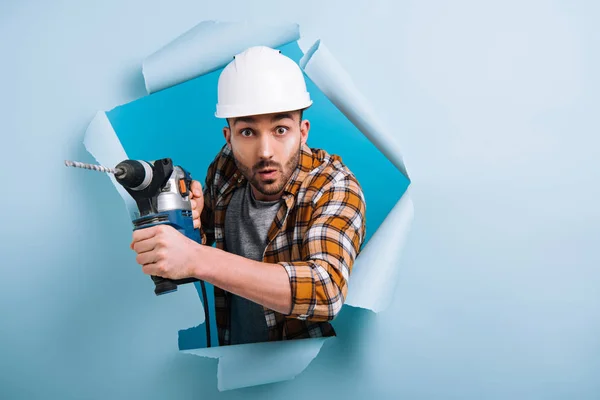 Reparador sorprendido en casco sosteniendo taladro eléctrico en papel roto, aislado en azul - foto de stock