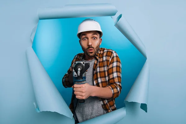 Schockierter Reparateur im Helm mit Bohrmaschine in zerrissenem Papier, auf blau — Stockfoto