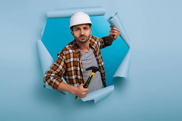Злой ремонтник в шлеме держит молоток в рваной бумаге, изолированный на голубом — стоковое фото