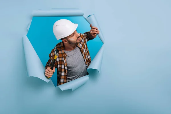 Aggressive worker in helmet holding hammer in torn paper, isolated on blue — Stock Photo