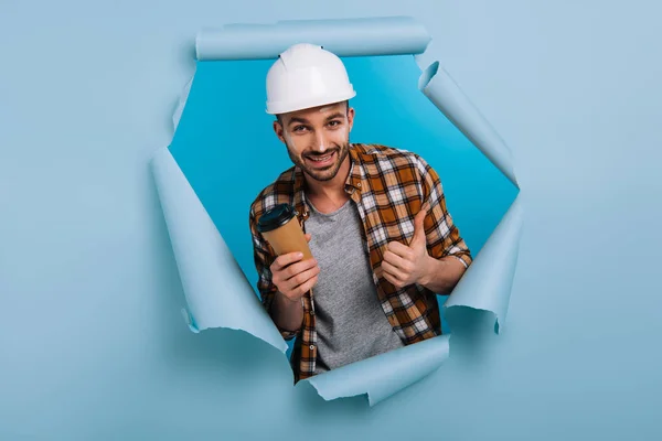 Счастливый ремонтник в шлеме, держащий кофе, чтобы пойти и показывающий большой палец вверх в разорванной бумаге, изолированной на голубом — стоковое фото