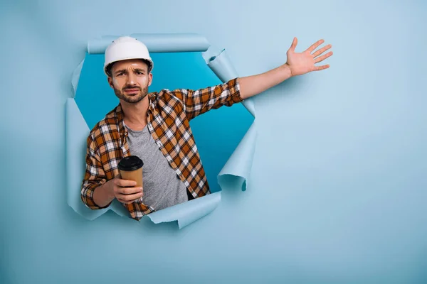 Frustrated foreman in helmet holding coffee to go in torn paper, isolated on blue — Stock Photo