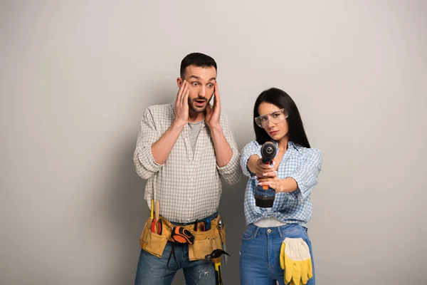 Uomo spaventato guardando operaia donna con trapano elettrico su grigio — Foto stock