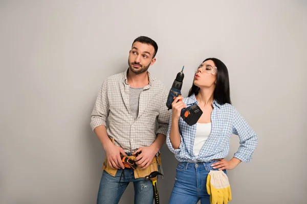 Skeptical repairman and confident female worker holding electric drill on grey — Stock Photo