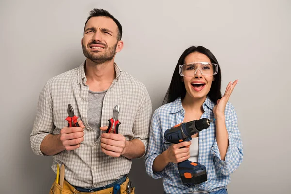 Stressed manual workers holding pliers and electric drill on grey — Stock Photo