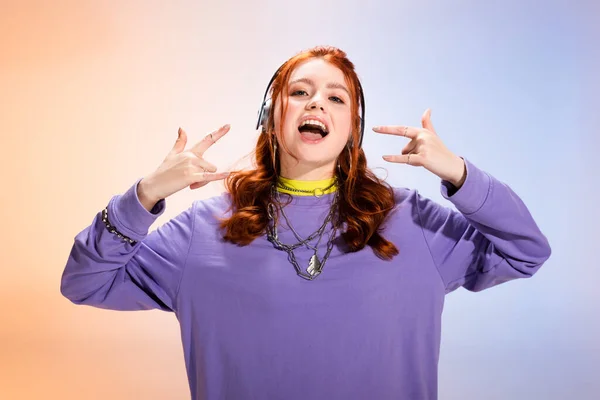Emotivo teen girl ascoltare musica con le cuffie e mostrando segni rock and roll, viola e beige — Foto stock