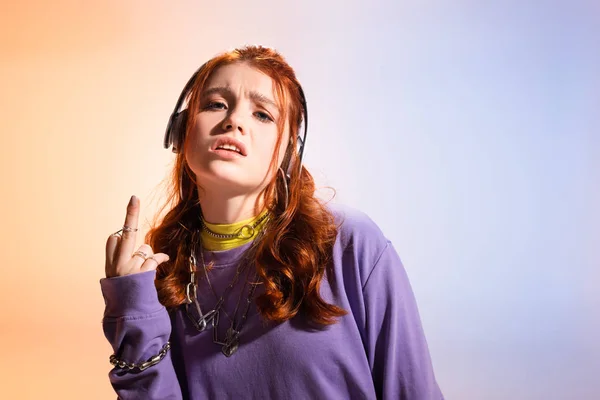 Angry teen girl listening music with headphones and showing middle finger, on purple and beige — Stock Photo