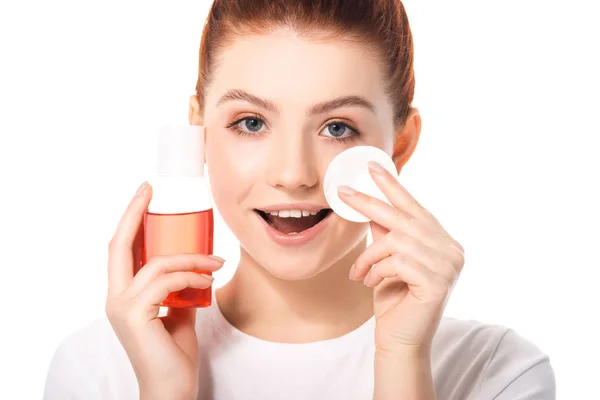 Adolescente excitado con piel limpia sosteniendo esponja de algodón y botella con removedor de maquillaje rojo, aislado en blanco - foto de stock