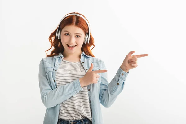Cheerful redhead teen girl listening music with headphones and pointing isolated on white — Stock Photo