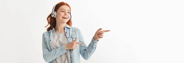 Панорамний знімок збудженого підлітка, який слухає музику з навушниками та вказує ізольовано на білому — стокове фото