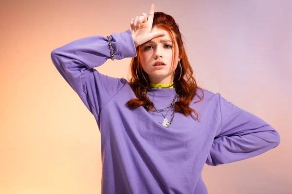 Redhead female teenager in bad mood showing loser sign, on purple and beige — Stock Photo