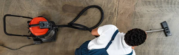 Top view of african american cleaner vacuuming floor in office, panoramic shot — Stock Photo