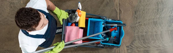 Top view of cleaner in uniform carrying cart with cleaning supplies, panoramic shot — Stock Photo