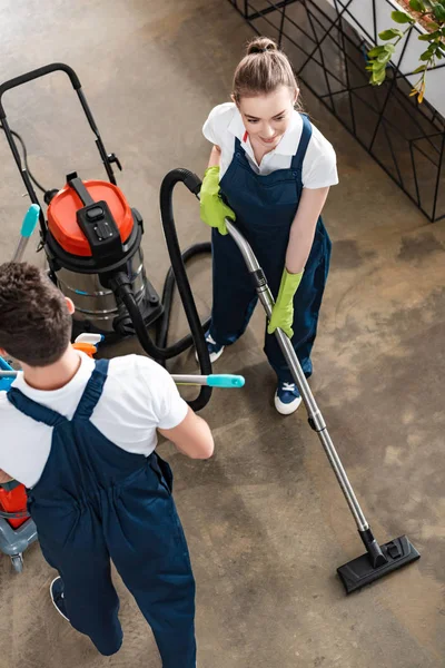 High angle view of attractive cleaner vacuuming floor near colleague carrying cart with cleaning supplies — Stock Photo