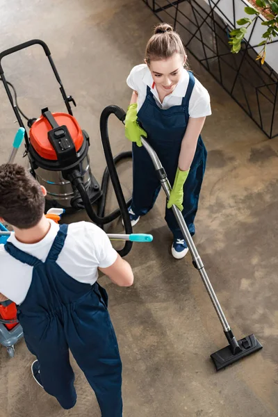 Vista de ángulo alto del piso de aspiración del limpiador atractivo cerca de colega que lleva el carro con los suministros de limpieza - foto de stock