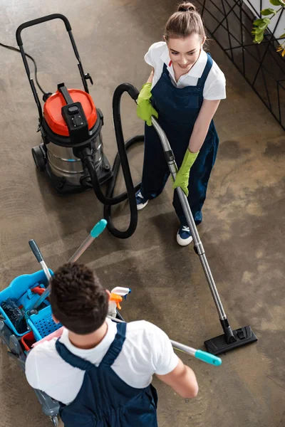 Visão de alto ângulo de chão aspirando aspirador de leite perto colega transportando carrinho com suprimentos de limpeza — Fotografia de Stock