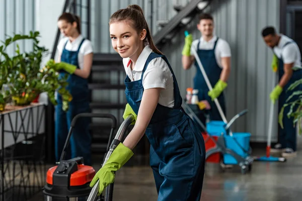 Smiling cleaner vacuuming floor in office near multicultural colleagues — Stock Photo