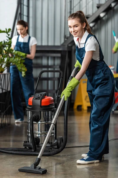 Enfoque selectivo del limpiador sonriente que aspira el piso cerca de las plantas de limpieza de colegas - foto de stock