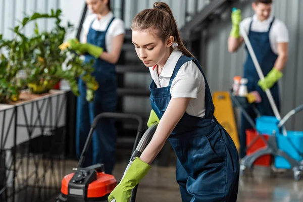 Selective focus of young cleaner vacuuming floor in office near colleagues — Stock Photo