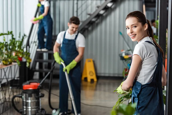 Attractive cleaner smiling at camera while colleagues cleaning office — Stock Photo