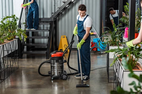 Young cleaner cleaning floor with vacuum cleaner near multicultural colleagues — Stock Photo