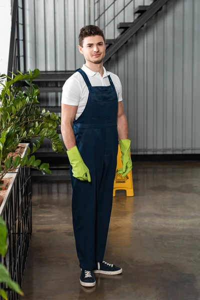 Lächelnde junge Putzfrau in Overalls und Gummihandschuhen blickt in die Kamera — Stockfoto