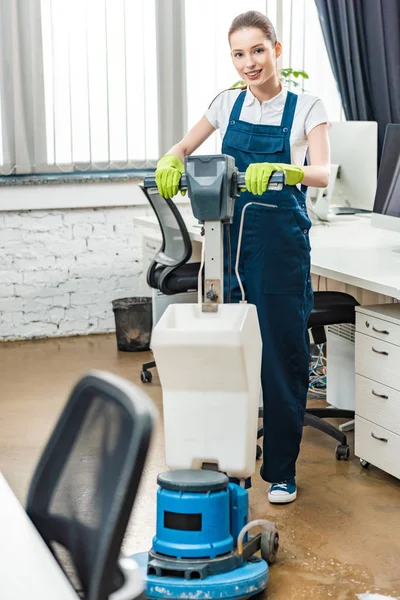 Limpiador sonriente mirando a la cámara mientras lava el piso con la máquina de limpieza - foto de stock