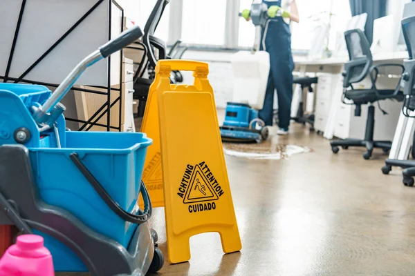 Selective focus of cart with buckets and wet floor caution sign, and cleaner washing floor with cleaning machine on background — Stock Photo