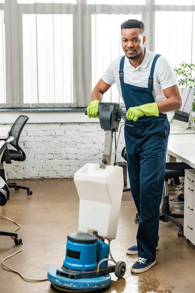 Sorrindo limpador da américa africana olhando para a câmera enquanto lava o chão com máquina de limpeza — Fotografia de Stock