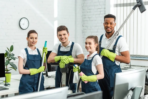 Limpiadores multiculturales felices mirando a la cámara mientras están de pie con los suministros de limpieza en la oficina - foto de stock