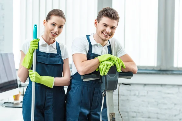 Two young cleaners in overalls smiling at camera while standing in office — Stock Photo