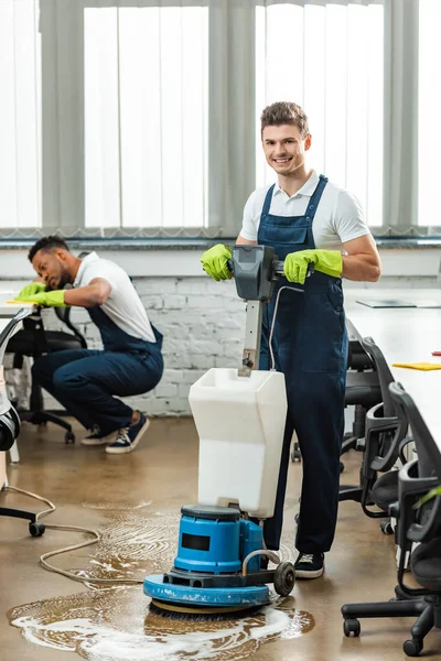 Happy cleaner washing floor with cleaning machine while colleague wiping desk — Stock Photo