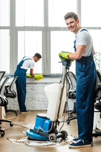 Limpiador sonriente lavando el piso con la máquina de limpieza cerca de colega limpiando ventana - foto de stock