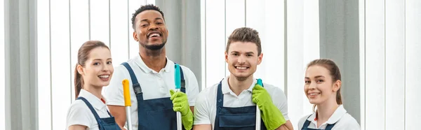 Tiro panorâmico de alegre equipe multicultural de limpadores olhando para a câmera enquanto estava no escritório — Fotografia de Stock