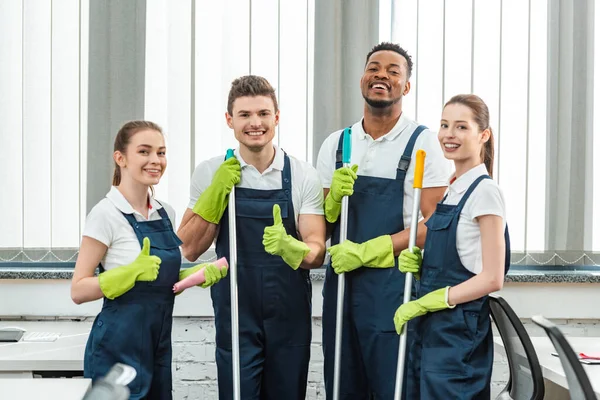 Equipe multicultural feliz de limpadores olhando para a câmera e mostrando polegares para cima — Fotografia de Stock