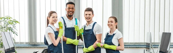 Prise de vue panoramique d'une équipe multiculturelle joyeuse de nettoyeurs regardant la caméra pendant qu'ils sont au bureau — Photo de stock