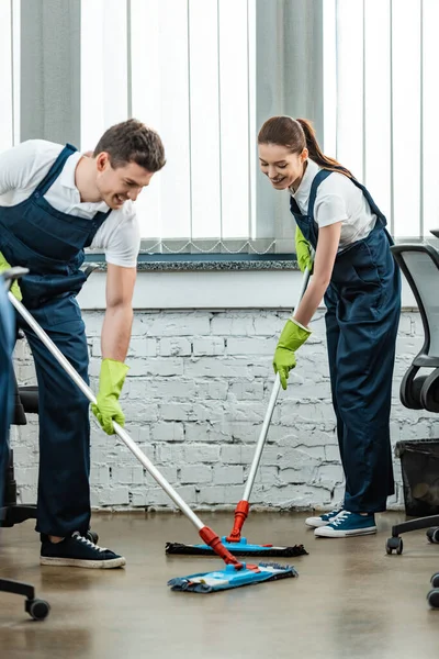 Zwei junge, gut gelaunte Putzfrauen in Uniform mit Wischmopp im Büro — Stockfoto