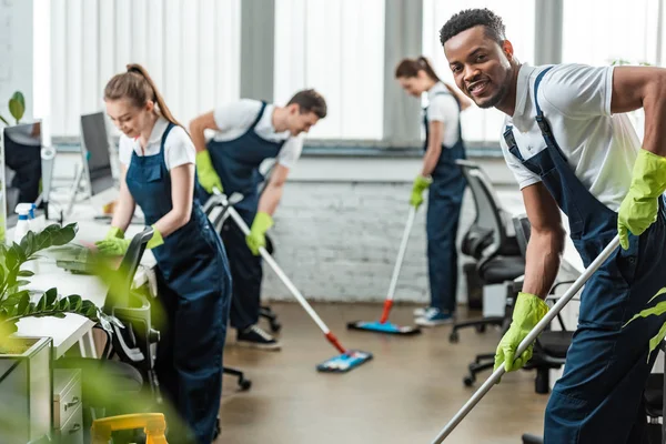 Foco seletivo de sorrindo equipe multicultural de limpadores lavando chão com esfregonas no escritório — Fotografia de Stock