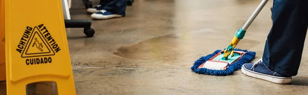 Cropped view of cleaner washing floor with mop near wet floor caution sign, panoramic shot — Stock Photo