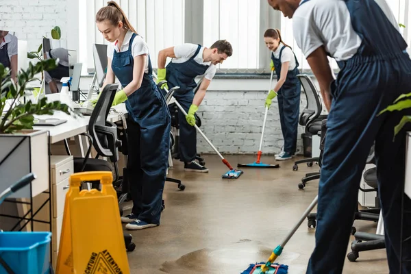 Equipo multicultural de jóvenes limpiadores lavando piso con fregonas en la oficina - foto de stock