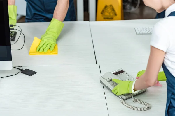 Vista parcial del limpiador limpiando el teléfono de la oficina cerca del escritorio de lavado colega - foto de stock