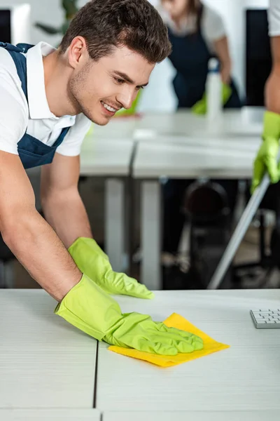 Limpiador joven y sonriente limpiando el escritorio de la oficina con trapo - foto de stock