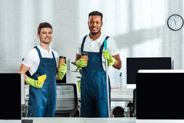 Smiling multicultural cleaners looking at camera while holding coffee to go in office — Stock Photo