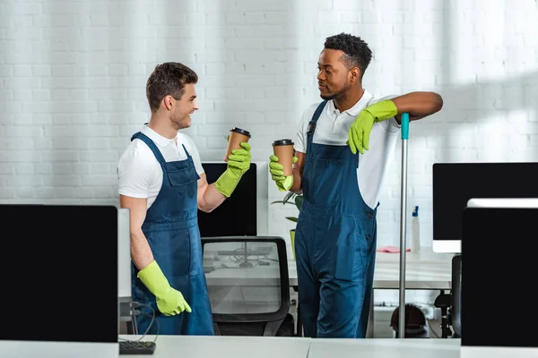 Two multicultural cleaners talking while holding coffee to go in office — Stock Photo