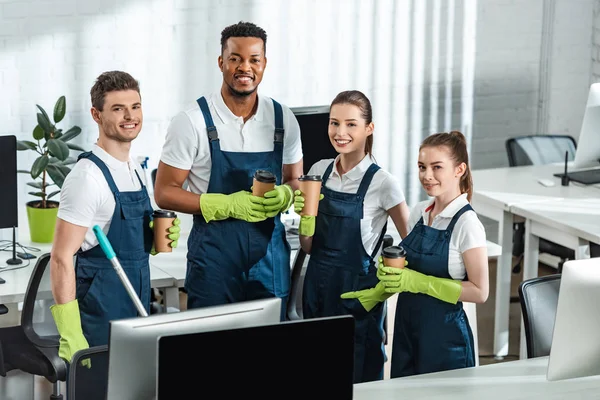 Limpiadores multiculturales alegres mirando a la cámara mientras sostiene el café para ir en la oficina - foto de stock