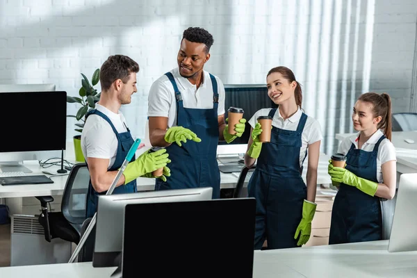 Happy multicultural cleaners talking while drinking coffee to go in office — Stock Photo