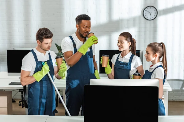 Cheerful multicultural cleaners drinking coffee to go in office — Stock Photo