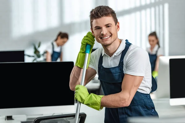 Apuesto, limpiador sonriente mirando a la cámara mientras está de pie cerca de los monitores de ordenador - foto de stock